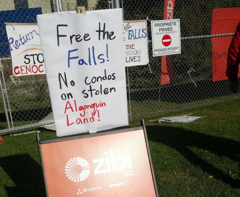 Photo by Vela Description: A handmade picket sign with the words "Free the Falls! No condos on stolen Algonquin Land!" sits directly on top of a corporate orange Zibi logo sign. 